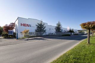 Das Bild zeigt das Firmengebäude der HENNgineered GmbH entlang einer Straße und vor blauem Himmel.