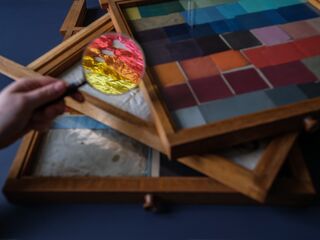 Vergrößerungsglas über bunten Farbmustern in Holzrahmen Eine Hand hält eine Lupe, die bunte Fäden in leuchtenden Farben zeigt, während Farbtafeln im Hintergrund sichtbar sind.
