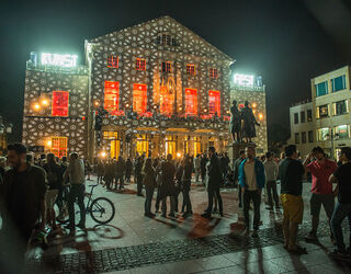 Eine lebhafte Menschenmenge versammelt sich vor dem kunstvoll beleuchteten Deutschen Nationaltheater Weimar mit roten Fenstern und einer Statue.
