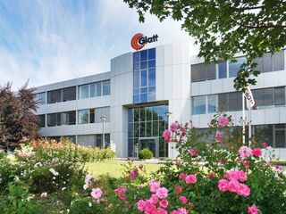 Glatt Unternehmenszentrale mit blühenden Gärten Ein modernes Firmengebäude mit einer Glasfassade, umgeben von bunten Rosen und grünen Bäumen unter einem blauen Himmel.