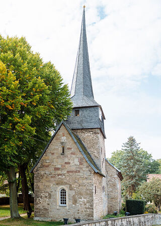 Eine kleine, steinerne Kirche in Gelmeroda mit einem hohen, spitzen Turm steht umgeben von grünen Bäumen und einem ruhigen Weg.