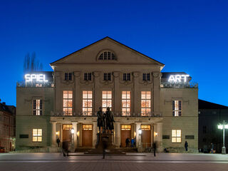 Die Fassade des Deutschen Nationaltheater Weimar wird von warmem Licht erhellt, während Passanten die Atmosphäre der Abenddämmerung genießen.