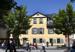 Schillers Wohnhaus Ein gelbes Gebäude mit grauen Fensterläden steht im Mittelpunkt, umgeben von grünen Bäumen und Passanten auf der Straße.