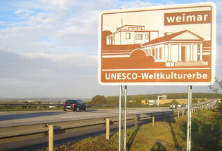 Autobahnschild UNESCO-Welterbe Weimar, Foto: Stefan Hopf, weimar GmbH Das Schild mit der Aufschrift 'Weimar' und 'UNESCO-Weltkulturerbe' steht an einer Autobahn, umgeben von sanften Hügeln.