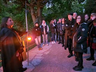 Eine Gruppe von Menschen hört einem Geschichtenerzähler zu, der eine Laterne in der Dämmerung hält.