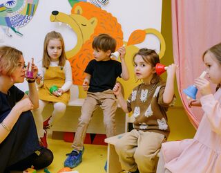 Symbolbild: Kinder mit Erzieherin, sitzen im Kreis, halten Spiel-Glöckchen und lauschen den Geräuschen, die sie machen
