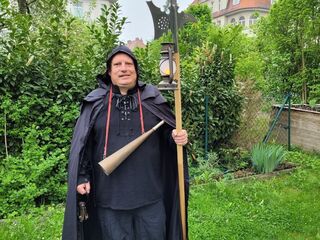 Ein Mann in einem schwarzen Umhang steht in einem Garten, hält eine Laterne in der Hand.