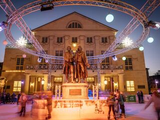 Eine festliche Szenerie mit einer Statue von Goethe und Schiller, umgeben von Lichtern und fröhlichen Menschen auf einer Eisbahn.