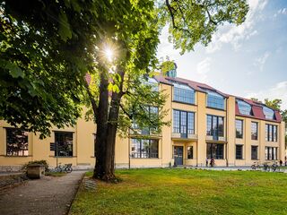 Das Gebäude mit seinen großen Fenstern und dem roten Dach wird von Sonnenlicht beleuchtet, während Menschen im Freien verweilen.