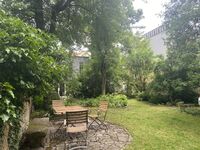 Ein idyllischer Garten mit einem runden Tisch und Stühlen, umgeben von üppigem Grün und einem modernen Gebäude im Hintergrund.
