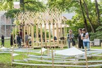 Eine Gruppe von Menschen steht in einem Park und betrachtet eine kreative Holzstruktur, umgeben von grünen Bäumen.