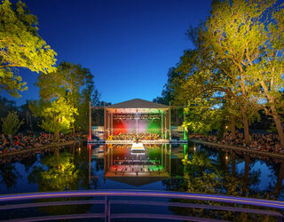 Die illuminierte Seebühne im Weimarhallenpark am Abend, umgeben von Bäumen, mit einem Publikum, das auf eine Aufführung am Wasser schaut.