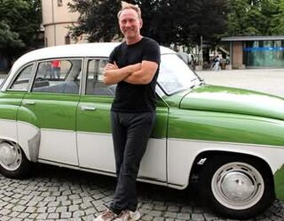 Grünes Oldtimer-Auto mit einem freundlichen Fahrer Der Fahrer posiert stolz vor seinem vintage Auto, das in einem lebhaften Grün und Weiß lackiert ist, umgeben von einer urbanen Kulisse.