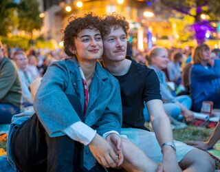 Paar genießt romantischen Abend bei Konzert im Freien Ein Paar sitzt eng umschlungen auf dem Boden, umgeben von einer fröhlichen Menschenmenge bei einem Konzert im Freien.