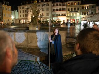 Nachtwächterführung Eine fröhliche Frau in einem Umhang steht vor dem Neptunbrunnen, umgeben von historischen Gebäuden und dem beleuchteten Markt.