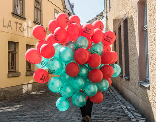 Eine Person trägt einen großen Haufen bunter Luftballons des Kunstfest Weimar in einer engen Gasse, umgeben von historischen Gebäuden.