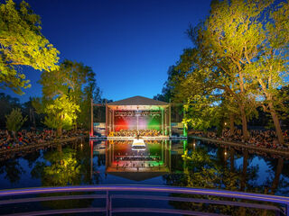 Die illuminierte Seebühne im Weimarhallenpark am Abend, umgeben von Bäumen, mit einem Publikum, das auf eine Aufführung am Wasser schaut.