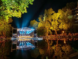 Eine lebhafte Konzertbühne erstrahlt nachts im Weimarhallenpark, umgeben von leuchtenden Bäumen und einem ruhigen Teich, der die Lichter reflektiert.