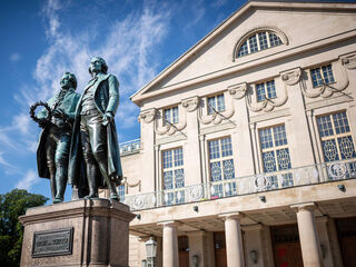Goethe- und Schiller-Denkmal auf dem Theaterplatz Goethe und Schiller, dargestellt in Bronze, halten einen Lorbeerkranz, während das Deutsche Nationaltheater Weimar im Hintergrund erstrahlt.