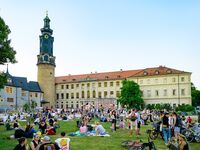Familien und Freunde genießen einen sonnigen Tag im Freien, während der alte Uhrturm und die Architektur im Hintergrund sichtbar sind.