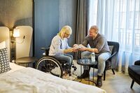 Eine Frau mit blonden Haaren im Rollstuhl und ein Mann tauschen sich in einem gemütlichen Hotelzimmer über ein Buch aus.