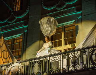 Eine Frau in einem eleganten weißen Kleid steht auf dem Balkon des Deutschen Naionaltheaters und hält einen großen, glänzenden Schirm über ihrem Kopf.