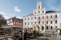Ein belebter Marktplatz mit bunten Ständen, umgeben von historischen Gebäuden und einer strahlend blauen Himmel.