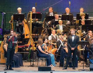 Ein unvergesslicher Abend voller Musik und Emotionen im Freien Ein charismatischer Moderator spricht auf der Bühne, während ein Orchester im Hintergrund bereit ist zu spielen.