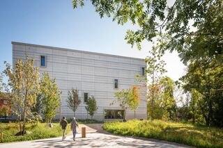 Bauhaus Museum Weimar Die Szenerie zeigt ein minimalistisches Gebäude, umgeben von grünen Bäumen und bunten Blumen, mit zwei Passanten.