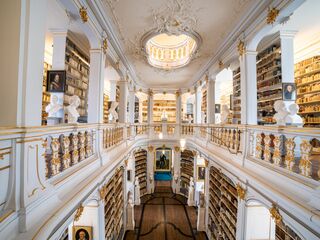 Anna Amalia Bibliothek Der Blick auf eine opulente Bibliothek mit goldenen Verzierungen, hohen Decken und einer zentralen Treppe, die zu weiteren Regalen führt.