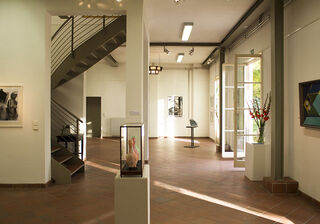 Ein heller Ausstellungsraum mit einer geschwungenen Treppe, Kunstwerken an den Wänden und einer offenen Tür zum Garten.