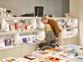 Museumsshop im Bauhaus-Museum Weimar, Foto: Florian Trykowski, TTG Ein Mann beugt sich über eine Frau im Rollstuhl, die ein Buch in einem hellen, einladenden Buchladen betrachtet, umgeben von vielen Titeln.