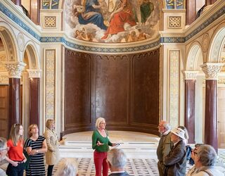 Führung durch die Schlosskapelle im Stadtschloss Eine Gruppe von Touristen hört einer Führerin in einem prächtigen Raum mit einem kunstvollen Wandgemälde zu.