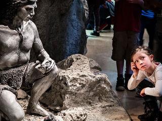 Museum fuer Ur- und Fruehgeschichte Ein prähistorischer Mann aus Stein sitzt auf einem Felsen, während ein kleines Mädchen mit einem nachdenklichen Ausdruck daneben steht.