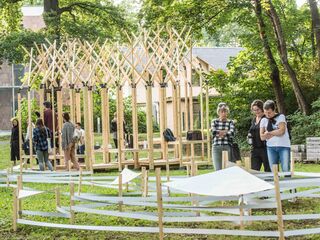 Eine Gruppe von Menschen steht in einem Park und betrachtet eine kreative Holzstruktur, umgeben von grünen Bäumen.