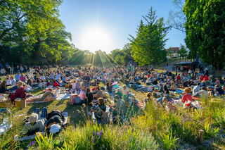 Konzertnacht Bella Italia Eine große Menschenmenge sitzt auf einer Wiese im Weimarhallenpark, umgeben von Bäumen, während die Sonne strahlend am Himmel aufgeht.