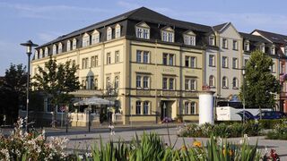 Außenaufnahme, Foto: Kaiserin Augusta Ein elegantes Hotelgebäude mit gelber Fassade, umgeben von blühenden Blumen und Bäumen, strahlt eine einladende Atmosphäre aus.