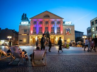 Der Theaterplatz ist voller Leben, mit Besuchern, die sich um ein kunstvoll beleuchtete Deutsche Nationaltheater Weimar und Liegestühle versammeln.