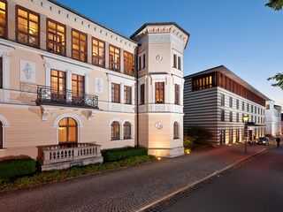 Eine ruhige Straße, gesäumt von Bäumen, führt zu einem beeindruckenden Gebäude mit einer Mischung aus traditionellem und modernem Design.