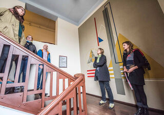 Fünf Menschen stehen in einem Treppenhaus und bewundern ein modernes Kunstwerk, das geometrische Formen und Farben kombiniert.