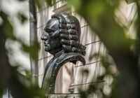 Eine bronzene Büste von Johann Sebastian Bach, die vor einer hellen Wand steht, umgeben von grünen Blättern.
