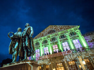 Im Vordergrund stehen Goethe und Schiller, während das Theater im Hintergrund mit lebhaften Lichtern und Projektionen erstrahlt.