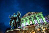 Im Vordergrund stehen Goethe und Schiller, während das Theater im Hintergrund mit lebhaften Lichtern und Projektionen erstrahlt.