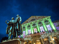 Im Vordergrund stehen Goethe und Schiller, während das Theater im Hintergrund mit lebhaften Lichtern und Projektionen erstrahlt.