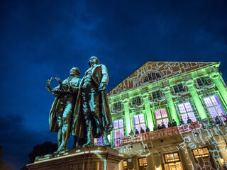 Im Vordergrund stehen Goethe und Schiller, während das Theater im Hintergrund mit lebhaften Lichtern und Projektionen erstrahlt.
