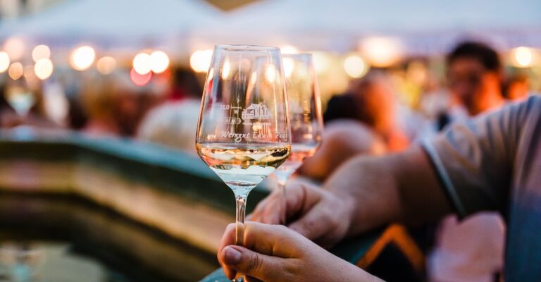 Im Vordergrund sind zwei Gläser mit Weißwein zu sehen, umgeben von einer lebhaften Menschenmenge und festlichen Ständen.