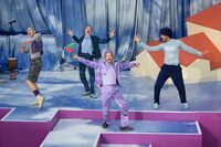 Vier fröhliche Darsteller in bunten Kostümen tanzen auf einer Bühne mit lila und blauen Hintergründen und einem Mikrofon.