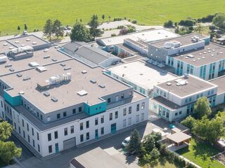 Moderne Architektur des Layertec Firmengebaeudes aus der Vogelperspektive In der Bildmitte ist ein weitläufiges, helles Gebäude zu sehen, das von grünen Flächen und Wohnhäusern umgeben ist.