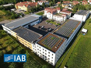 Luftaufnahme des Instituts für Angewandte Bauforschung Weimar gGmbH Eine Luftaufnahme eines modernen Gebäudekomplexes mit Solarpanelen auf den Dächern und einer grünen Umgebung.
