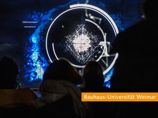 Interaktive Kunstinstallation an der Bauhaus-Universität Weimar Eine Gruppe von Menschen betrachtet eine digitale Präsentation mit abstrakten, leuchtenden Grafiken und Zahlen auf einem großen Bildschirm.